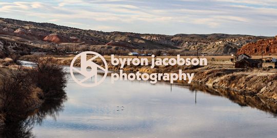 The Byron Jacobson Photography logo placed on a Wyoming river with a view of a townscape